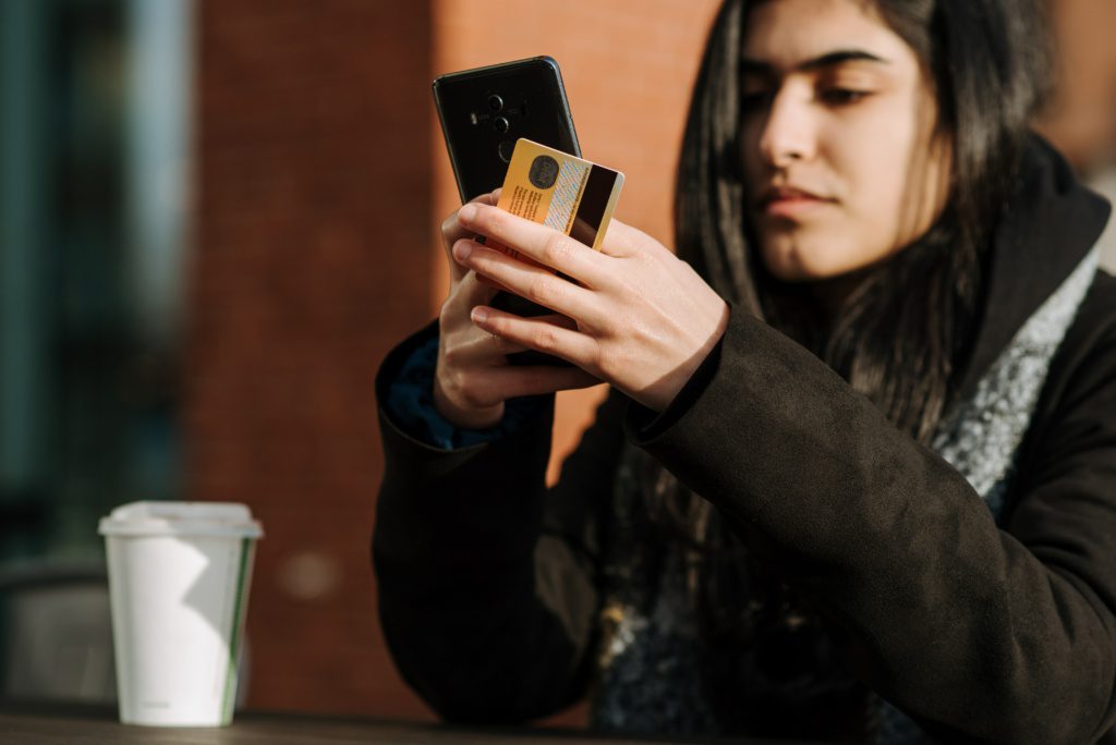 Mulher usando o celular e segurando um cartão pesquisando sobre o juros do cartão de crédito